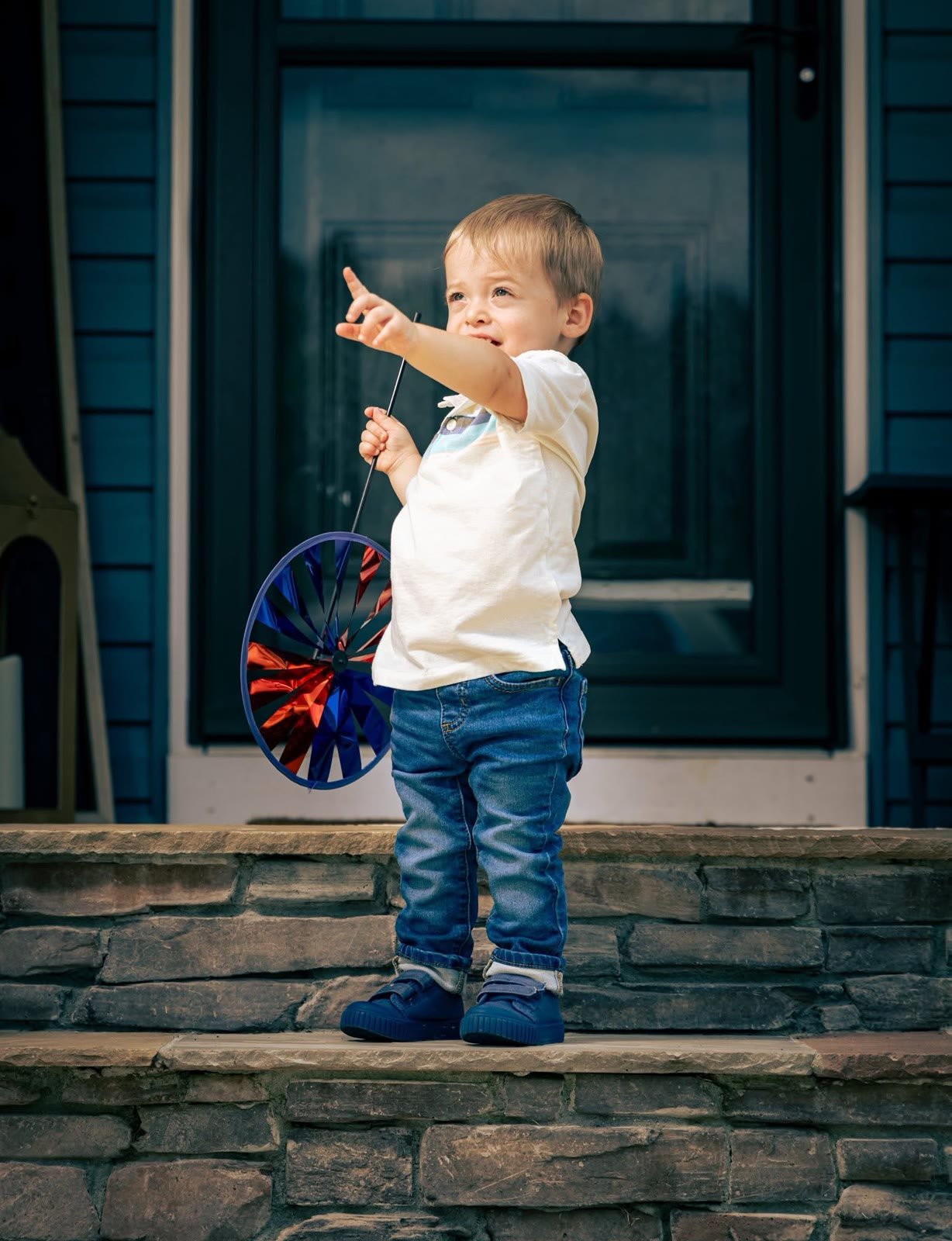 Toddler holding a red and blue pinwheel on stone porch steps, pointing at something off camera
