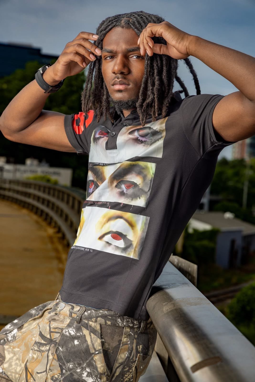 Man with long locs leaning on an overpass rail, hands in his hair, city behind him