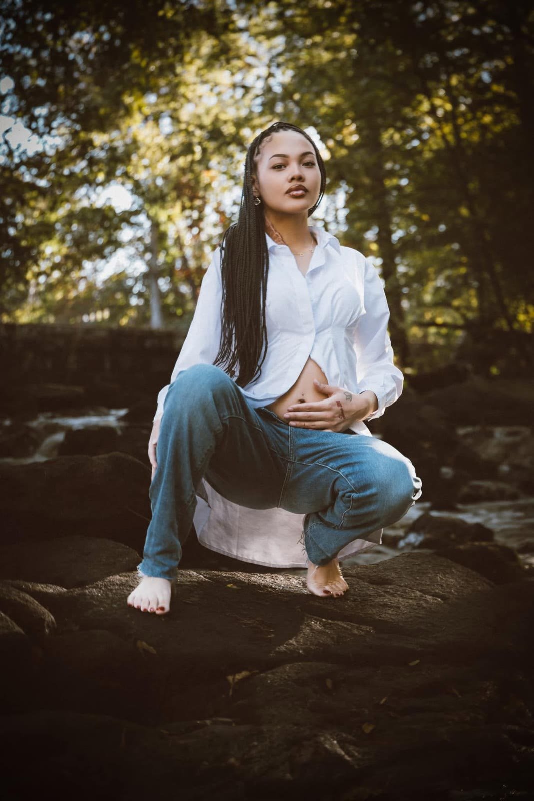 Woman in a white shirt and jeans crouching barefoot on creek rocks under autumn trees