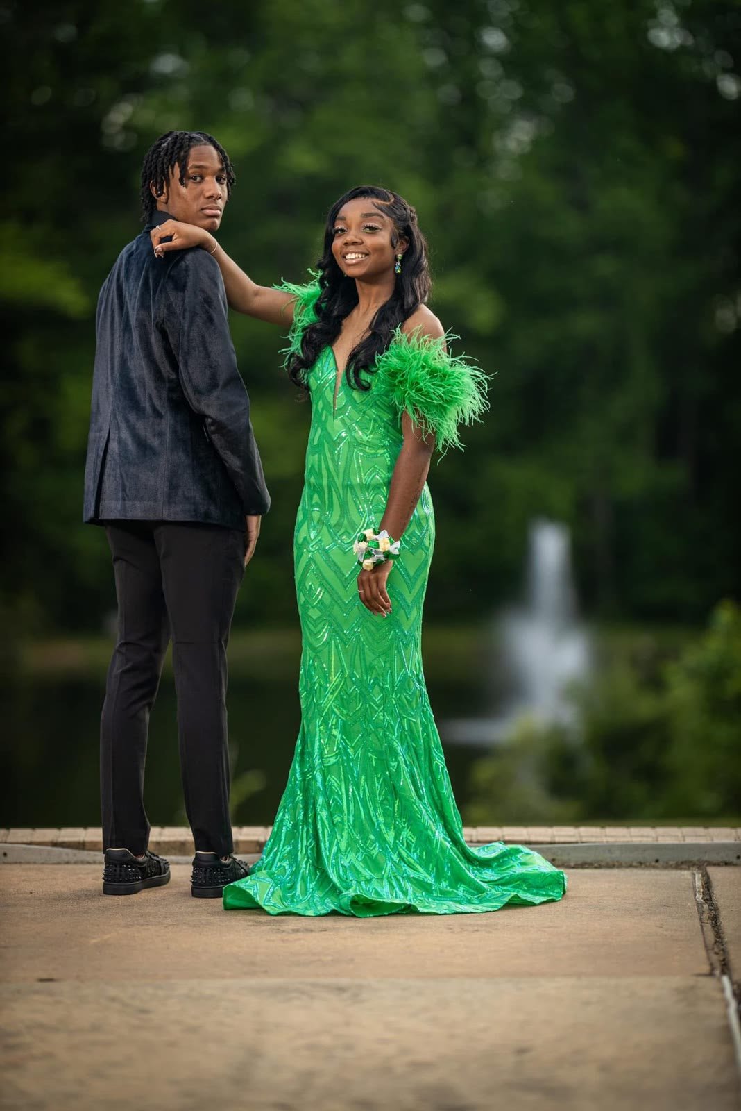 Prom couple by a park fountain, she in a green sequin gown with feather sleeves, he in a velvet jacket