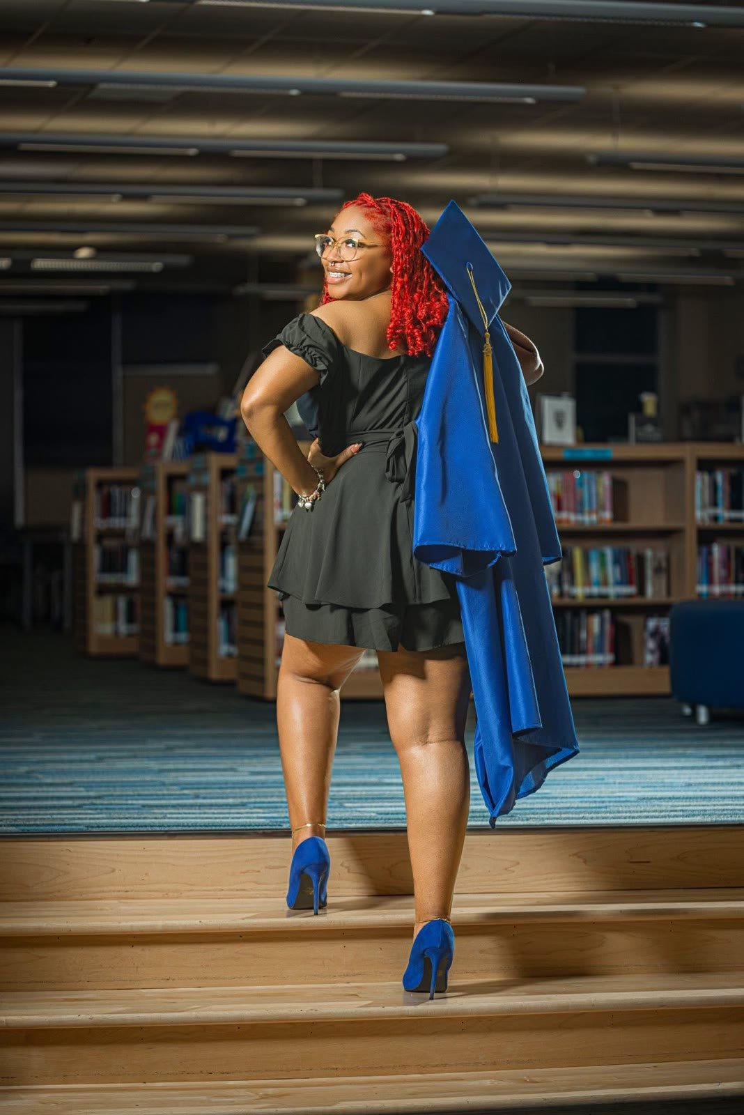 Graduate in a black dress and blue heels on library steps, gown over her shoulder, smiling back at the camera